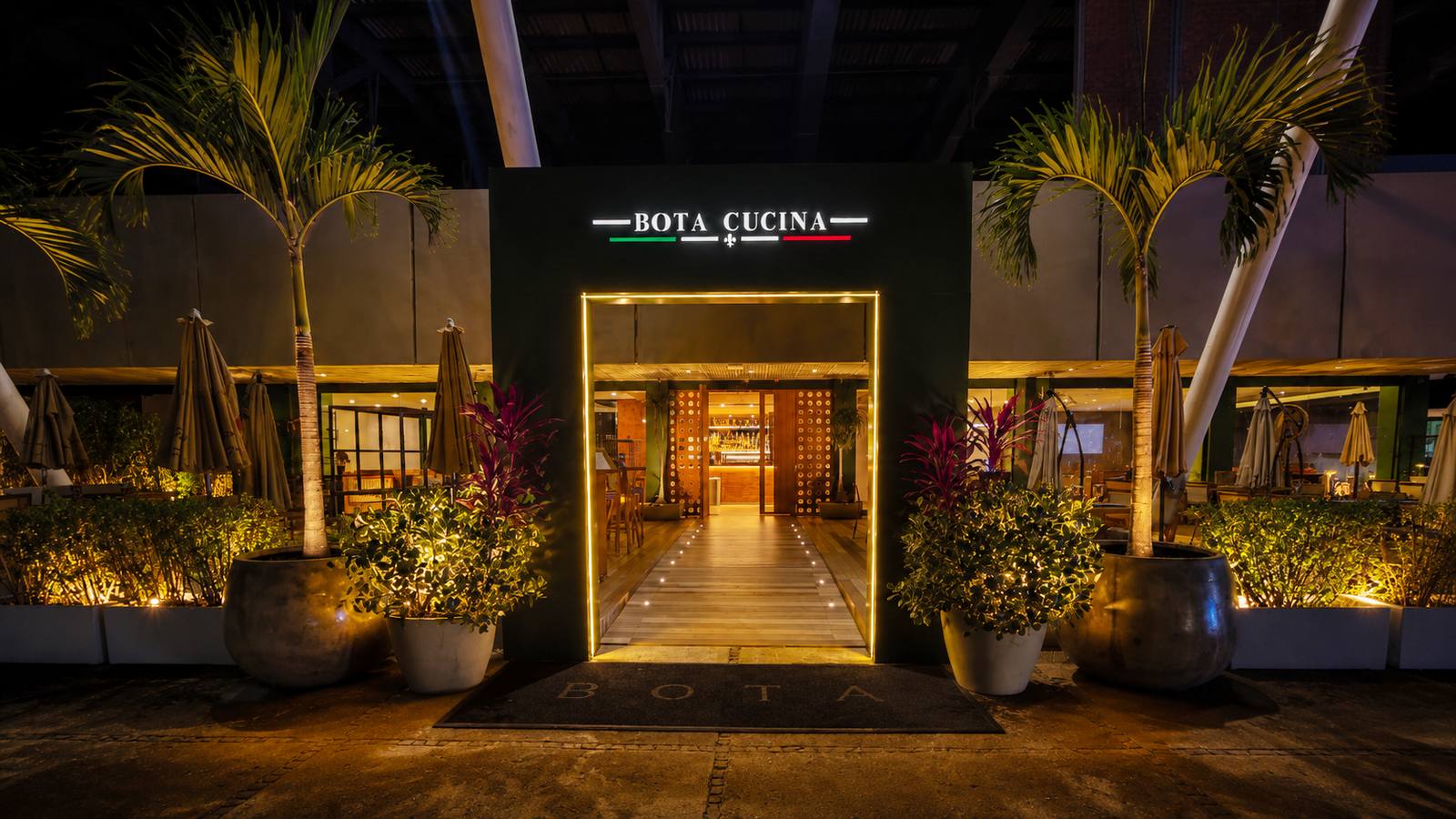 Entrada iluminada do BOTA Cucina à noite, com o letreiro BOTA CUCINA